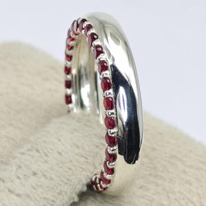 May include: A polished silver ring featuring a single row of small, round, deep red rubies set along one side of the band. The metal surface is smooth and reflective, contrasting with the vibrant, faceted texture of the embedded gemstones.
