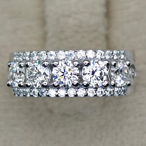 Könnte beinhalten: Ein silberner Ring mit mehreren Reihen funkelnder, klarer Edelsteine. Der Ring hat eine zentrale Reihe größerer Steine, flankiert von kleineren Steinen, die ein schillerndes Aussehen erzeugen. Das Design ist symmetrisch und elegant.