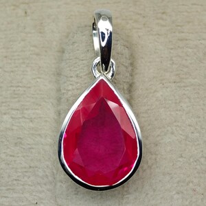 May include: A teardrop-shaped pendant with a vibrant red gemstone set in a polished silver frame. The gemstone has multiple facets, reflecting light. The pendant has a silver bail for attaching to a chain.