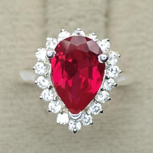 May include: A silver ring featuring a large, pear-shaped, deep red gemstone at the centre. The central stone is surrounded by a halo of small, round, clear, sparkling stones set in a scalloped pattern, creating a bright, contrasting frame.
