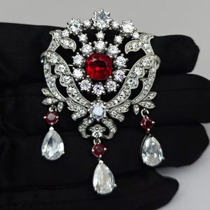 Könnte beinhalten: Eine verzierte silberne Brosche mit einem großen, quadratischen, roten Edelstein in der Mitte, umgeben von kleineren, klaren Steinen. Die Brosche hat ein florales Design mit hängenden tropfenförmigen, klaren Steinen und kleineren roten Steinen.