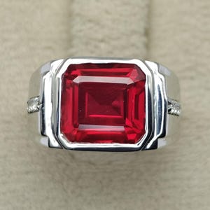 May include: A silver-toned ring featuring a large, rectangular, faceted red gemstone set in the centre. The band has a wide, polished metal design with small, clear, sparkling stones embedded along the sides of the shank near the setting.