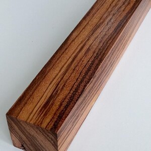 May include: A rectangular wooden block featuring a distinct grain pattern with alternating light and dark brown stripes. The piece has a smooth, polished finish and sharp, clean edges, showcasing the natural texture and color variations of the wood.