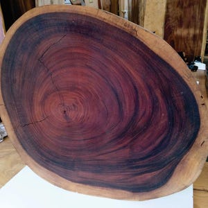 May include: A large, circular wood slab featuring deep reddish-brown and dark charcoal grain patterns with a natural bark edge. The surface displays visible growth rings and a crack. Text on the slab reads 25.75 x 22.25 x 2.25 inches.
