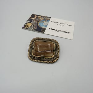 May include: A square-shaped brooch with a brown and white agate stone set in a gold-toned metal frame. The frame has a decorative design.