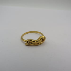 May include: A gold-colored ring featuring a frog design. The frog has two small, round accents, possibly gemstones, for eyes. The ring band is thin and simple, and the frog is the focal point.