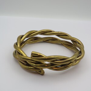 May include: A gold-coloured metal bracelet with a woven design. The bracelet is circular, with an open end. The metal has a slightly aged appearance. The bracelet is a piece of jewellery.