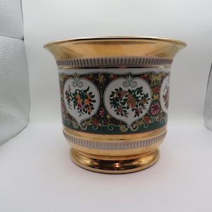 Peut inclure: Un pot de fleurs en céramique de couleur or, avec un large bord et une base en piédestal. Le pot présente une bande décorative avec des motifs floraux dans les tons de rose, vert et jaune. Le bord et la base ont une finition dorée.