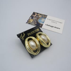 May include: A pair of gold tone clip-on earrings with faux pearl accents. The earrings are on a black card with the text "First Fashion" printed on it.