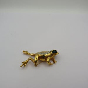 May include: A gold-coloured frog brooch with clear rhinestone detailing along its back and dark green eyes. The frog is depicted crawling, with its legs extended. The brooch has a pin fastening on the reverse.