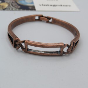 May include: A copper-toned bangle bracelet with a rectangular cutout in the centre. The bracelet has a simple, minimalist design.