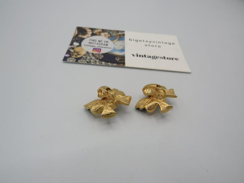 May include: A pair of gold-tone bow-shaped clip-on earrings. The earrings are made of metal and have a textured surface.