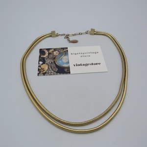 May include: A vintage gold-tone necklace with a double-strand design. The necklace features a textured, snake-like chain and a simple clasp. The necklace is displayed on a white surface with a vintage store tag.