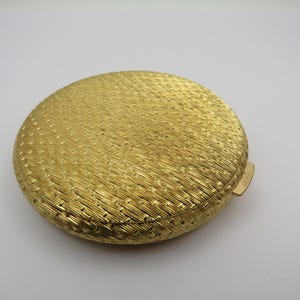 Puede incluir: Un estuche compacto redondo de color dorado con un patrón texturizado y tejido. El estuche compacto tiene un pequeño cierre rectangular en el lateral. La superficie está cubierta con un patrón repetido de líneas y puntos.