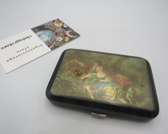 May include: A rectangular black case with a hinged lid, featuring a painted scene of figures in a garden. The artwork depicts a woman in a blue dress and a man in a red robe. A business card with the text "bigetsvintage store" is also present.