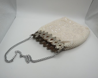 May include: A cream-colored beaded handbag featuring a textured surface with intricate floral patterns. The bag is secured at the top with a silver-toned metal accordion-style frame and includes a matching silver-toned metal chain shoulder strap.