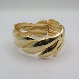 May include: Gold-coloured bracelet with a leaf design. The hinged bracelet features multiple overlapping leaf shapes, creating a textured and elegant look. The bracelet is made of a shiny, polished metal.