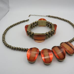 May include: A jewellery set featuring a necklace and bracelet. The necklace and bracelet have a bronze-coloured beaded chain. The necklace has large, orange, faceted pendants. The bracelet has orange rectangular accents.