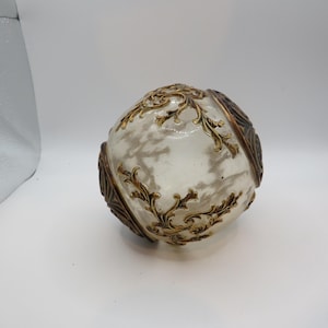 May include: A spherical glass object featuring a translucent white and grey marbled finish. The surface is adorned with raised, metallic gold-toned filigree scrollwork patterns that wrap around the sides, creating a textured and ornate appearance.