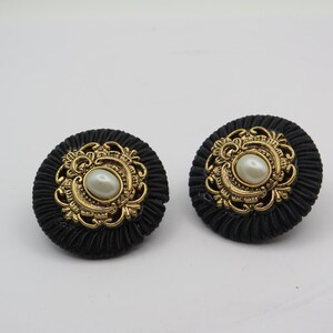 K&ouml;nnte beinhalten: Paar schwarze Knopf-Ohrringe mit goldfarbenem Filigran und einer zentralen Kunstperle. Die Ohrringe haben ein strukturiertes, kreisf&ouml;rmiges Design mit einem goldfarbenen, verzierten Rahmen um die Perle. Auf wei&szlig;em Hintergrund.