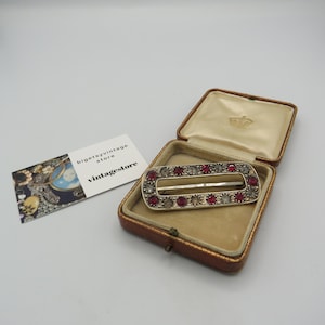 Peut inclure: Une broche rectangulaire en argent et rouge sertie de pierres rouges et claires dans un écrin doublé de velours. La broche a une ouverture centrale.