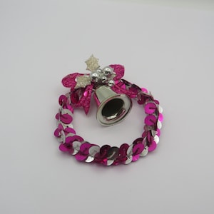May include: A festive Christmas wreath ornament with a silver bell, pink sequins, and a pink bow. The wreath is circular and adorned with shiny pink and silver sequins. The bell is silver and has a small clapper.