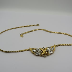 May include: A gold-toned necklace with a woven chain and a central pendant. The pendant features a cluster of clear, faceted crystals set in a decorative gold-tone frame. The necklace is displayed on a white surface.