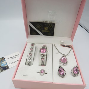 Pode incluir: Um conjunto de joias rosa numa caixa rosa. O conjunto inclui um relógio, uma pulseira, um anel, um colar e brincos. O relógio e a pulseira têm pedras rosa. O colar e os brincos têm formato de gota.