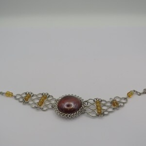 Könnte beinhalten: Silberfarbenes Armband mit einem großen, ovalen, rotbraunen Mittelstein, umgeben von einem dekorativen Silberrahmen. Das Armband hat ein filigranes, offenes Design mit bernsteinfarbenen Perlen und einem Verschluss.