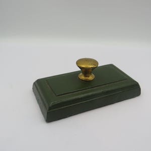 May include: A vintage ink blotter. It has a dark green rectangular base and a gold-coloured knob. This classic office supply would have been used to absorb excess ink from handwritten documents.