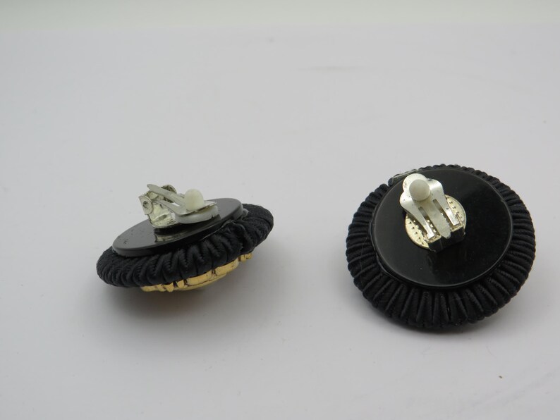 K&ouml;nnte beinhalten: Paar schwarze Clip-Ohrringe mit rundem Design. Die Ohrringe haben einen schwarzen, strukturierten Rand, eine schwarze Mitte und goldfarbene Akzente. Der Clip-Mechanismus ist silberfarben.