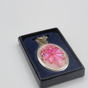 Könnte beinhalten: Eine Halskette mit einem rosa Blumenanhänger in einer schwarzen Samtbox. Der Anhänger ist oval geformt und zeigt eine rosa Blume mit einem Perlenmittelpunkt.