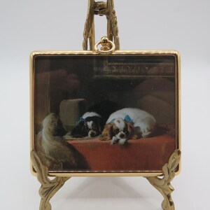 May include: A square framed print featuring a painting of three spaniel dogs resting on a red surface, with two dogs wearing blue bows. The artwork is encased in a thin gold-toned metal frame with a small top loop, displayed on a gold-toned stand.