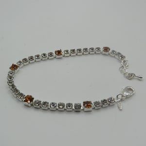 May include: A silver-tone tennis bracelet featuring a series of clear, round-cut stones interspersed with four larger, square-cut amber-coloured stones. The bracelet includes a lobster claw clasp, an adjustable chain extension, and a small metal tag.