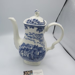 May include: A white ceramic teapot with a blue and white floral pattern. The teapot has a lid and a handle. The pattern features a cottage and trees.