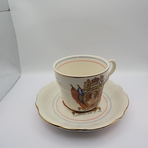 Op de afbeelding: Crèmekleurige theekop en schotel set. De kop en schotel hebben een gouden rand en een ontwerp met een kroon, vlaggen en een portret. De schotel heeft de tekst "Coronation June 2nd 1953".