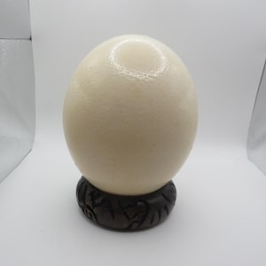 May include: A large, smooth, off-white ostrich egg resting securely on a dark brown, circular wooden stand. The egg features a natural, slightly textured surface with subtle variations in color, while the base displays intricate carvings.