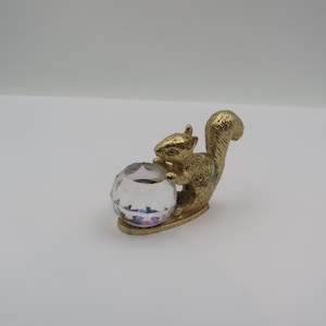 May include: A gold-colored squirrel figurine holding a faceted crystal ball. The squirrel is detailed with textured fur and a bushy tail. The crystal ball has a clear, multi-faceted design. The figurine is on a white background.