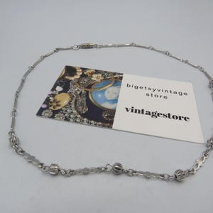 May include: A silver chain necklace with decorative ball charms. The necklace is on a white surface with a business card that reads "bigetsyvintage store" and "vintagestore".