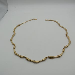 Könnte beinhalten: Eine goldfarbene Halskette bestehend aus einer Reihe von texturierten, zylindrischen Gliedern. Die Kette verfügt über ein kurzes Verlängerungsstück mit einem Hakenverschluss am Ende, was ein durchgehendes, flexibles Design mit metallischem Glanz ergibt.