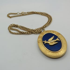 Pode incluir: Um colar com pendente oval em tom dourado com um centro de esmalte azul profundo e um emblema de pássaro em relevo. O pendente é rodeado por uma borda dourada texturizada com caracteres chineses gravados e uma corrente dourada a condizer.