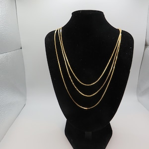 Puede incluir: Un collar de cadena de oro de múltiples hilos que presenta tres cadenas delgadas y delicadas de diferentes longitudes que caen en capas. El acabado dorado metálico refleja la luz, resaltando los eslabones finos y uniformes de cada cadena.