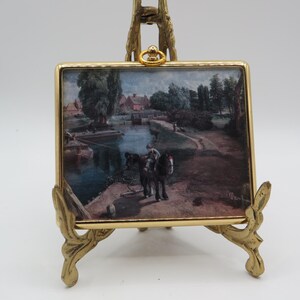May include: A rectangular decorative plaque featuring a landscape painting of a canal scene with a horse and rider, encased in a polished gold-tone metal frame. The plaque is supported by an ornate, matching gold-tone metal display stand.