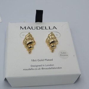 May include: Gold-plated earrings shaped like seashells with cubic zirconia accents. The earrings are displayed on a white box with the brand name "MAUDELLA" and the text "18ct Gold Plated. Designed in London. maudella.co.uk @maudellalondon".