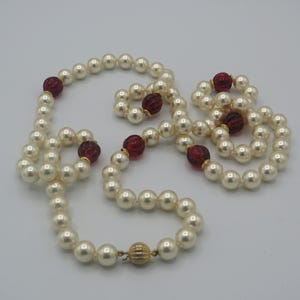 May include: A cream-colored pearl necklace with red glass beads interspersed throughout. The necklace has a gold-colored clasp and is displayed on a white surface. The pearls are round and evenly sized, creating a classic jewelry piece.