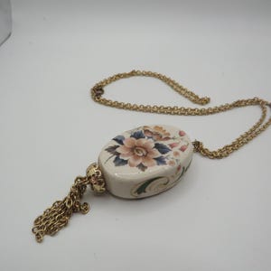 Puede incluir: Un collar de cadena en tono dorado con un colgante de cerámica de forma ovalada. El colgante es de color crema con un diseño floral pintado en tonos rosa, azul y verde. Una tapa de metal dorado sujeta una borla de cadena dorada al colgante.
