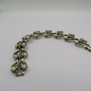May include: Silver-toned bracelet featuring a leaf and pearl design. The bracelet has a series of stylized leaves, each adorned with small, round, off-white pearls. The clasp is a simple, secure design. A delicate piece of jewelry.