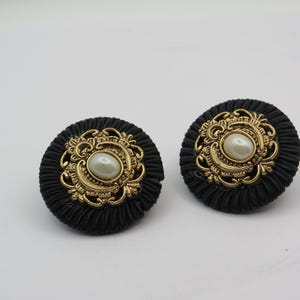 K&ouml;nnte beinhalten: Ein Paar schwarze Ohrringe mit goldenen Verzierungen und einer zentralen perlenartigen Perle. Die Ohrringe haben ein strukturiertes, kreisf&ouml;rmiges Design und sind vor wei&szlig;em Hintergrund platziert.