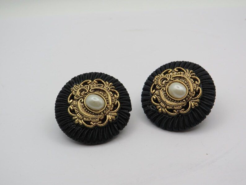 K&ouml;nnte beinhalten: Ein Paar schwarze Ohrringe mit goldenen Verzierungen und einer zentralen perlenartigen Perle. Die Ohrringe haben ein strukturiertes, kreisf&ouml;rmiges Design und sind vor wei&szlig;em Hintergrund platziert.