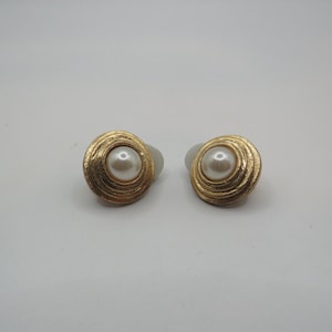 May include: A pair of round clip-on earrings featuring a central white faux pearl surrounded by a textured gold-tone metal frame with concentric circular ridges. The design is symmetrical, showcasing a polished finish on the pearl and a matte, brushed appearance on the metallic outer edge of each earring.